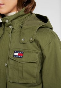 Zbliżenie na osobę w oliwkowej zielonej kurtce z przednią kieszenią i naszywką z logo Tommy Jeans na piersi.