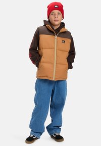 En pojke som bär en röd mössa, brun huvtröja, beige och brun zip-up väst, lösa blå jeans och svarta sneakers, står med händerna i fickorna.