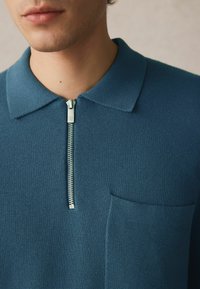 Blå strikket polo skjorte med kort lynlås, krave og en frontlomme. Teksturen er ribbet, hvilket giver et glat, tætsiddende udseende.