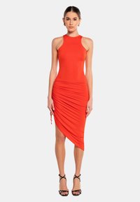 Rotes ärmelloses Kleid mit enganliegendem Oberteil, asymmetrischem Saum und gerafften Details auf einer Seite. Getragen mit schwarzen Riemchen-Heels.