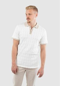Polo shirt blanc à manches courtes avec un motif texturé, col plat en tissé beige, patte de boutonnage à deux boutons, associé à un pantalon de couleur claire.