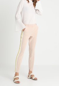 Světle růžové tailored kalhoty s bílými a žlutými bočními pruhy, v kombinaci s bílým topem s dlouhými rukávy a sklady na zvonových rukávech.