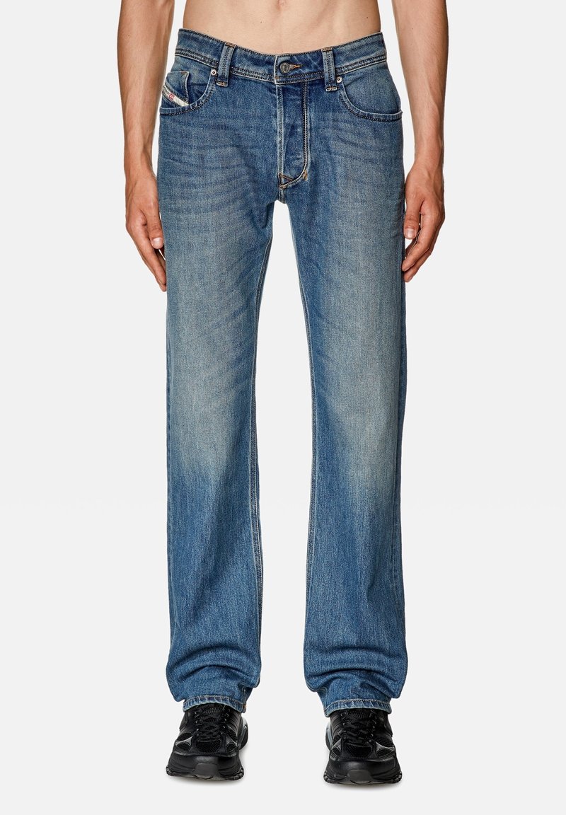 Blå denimjeans med rak bendesign, blekt tvätt, fem fickor och knappgylf. Bäraren har på sig svarta sneakers.