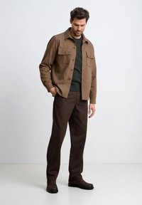 Brązowa teksturowana kurtka koszulowa z dwoma kieszeniami na piersi, noszona na zielonym swetrze i brązowych spodniach, zestawiona z ciemnobrązowymi butami.