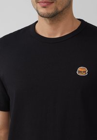 Czarny bawełniany t-shirt z małym haftowanym logo burgera na lewej piersi. Okrągły dekolt i krótkie rękawy.