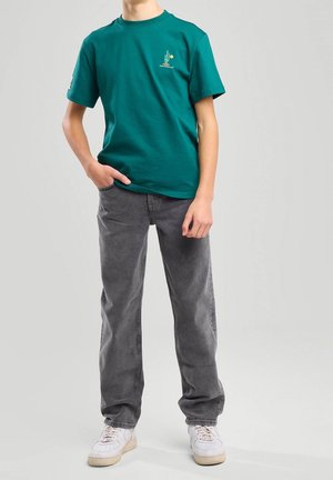 Henkilö, jolla on päällään teelasin turkoosi graafinen t-paita, harmaat suoralahkeiset farkut ja valkoiset tennarit, seisoo yhdellä kädellä taskussa yksinkertaisella taustalla.