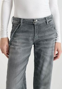 Jean en denim gris avec une coupe droite, doté d'une fermeture boutonnée et zippée standard. Coutures visibles et poches avant sont présentes.