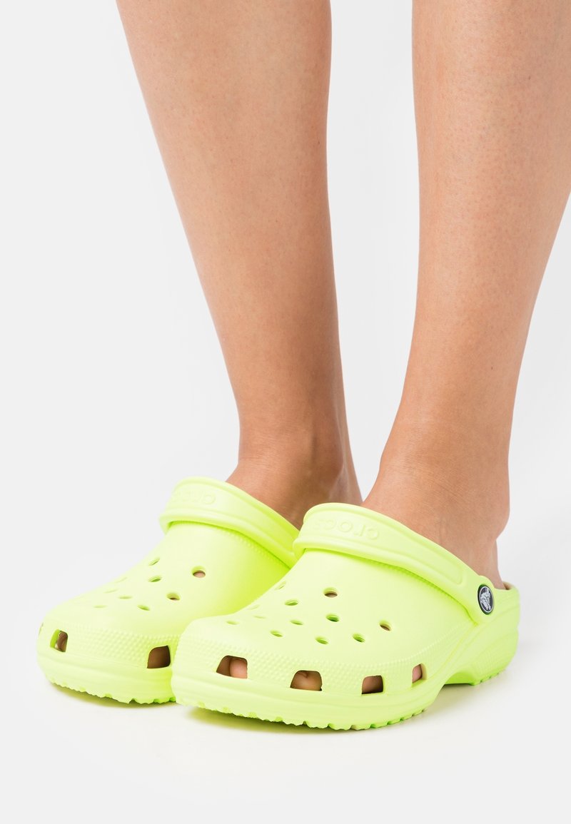 Pieds portant des sabots Crocs jaune néon vif avec des trous de ventilation et des brides de talon réglables sur fond blanc.