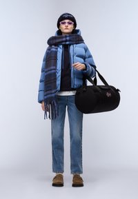 Giacca a vento azzurra chiara, sciarpa blu navy con righe, maglione scuro e jeans blu sbiaditi. Borsa a tracolla nera con dettagli a toppe e scarpe marroni.