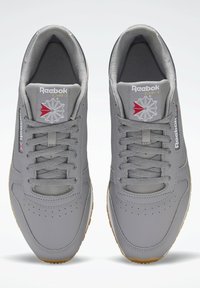 Pelēki Reebok sporta apavi ar gludu ādas virsmu, apaļu purngalu un perforētiem elementiem. Aprīkoti ar gumijas zoli un izšūtiem logotipa akcentiem.