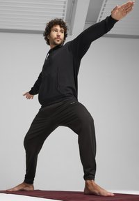 Haut à capuche noir avec poche avant et cordon de serrage, associé à un pantalon noir fuselé ; les deux en tissu lisse et flexible, adaptés pour le yoga.