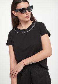 T-shirt nera a maniche corte con scollatura rotonda caratterizzata da ricamo di testo bianco. Il modello indossa occhiali da sole a forma di occhio di gatto, che esaltano il contrasto dell'outfit.