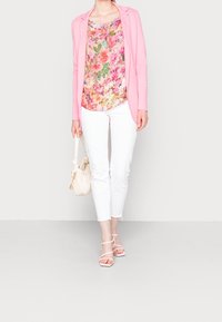 Blazer rosa, top floral multicolor, pantalones blancos ajustados y sandalias de tira rosas pálidas. Sosteniendo un bolso blanco con textura plizada.