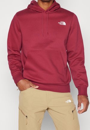 Mężczyzna ubrany w bordową bluzę z kapturem The North Face i beżowe spodnie z logo The North Face na klatce piersiowej i udzie, ręce rozluźnione wzdłuż ciała.