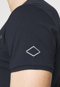 Mörkblå t-shirt med rund halsringning och korta ärmar, med en vit diamantlogotyp broderad på vänster ärm.