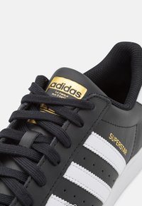 Svart sneakers i läder med vita ränder, gulddetaljer på varumärkeslogotypen och texten "SUPERSTAR". Har runda skosnören och perforerade detaljer.