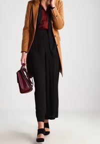 Abrigo largo marrón sobre una blusa burdeos y pantalones de pierna ancha negros, combinados con tacones negros y un bolso burdeos. Materiales lisos y estructurados.