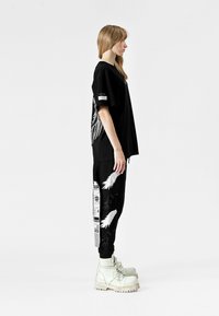 Camiseta negra de gran tamaño con estampado gráfico en blanco, combinada con pantalones de chándal negros que presentan diseños de alas en blanco y acentos gráficos. Botas blancas.