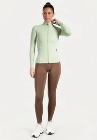 Hellgrüne, taillierte Reißverschluss-Jacke mit langen Ärmeln, kombiniert mit braunen Leggings und weißen Turnschuhen, mit glatter Textur und minimaler Gestaltung.
