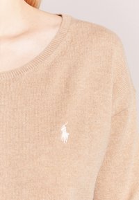 Beżowy sweter z dzianiny z ribbowym kołnierzykiem oraz małym białym logo haftowanym na piersi. Miękka tekstura, luźny krój.