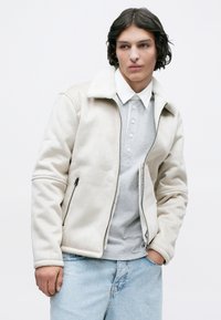 Giacca in finta shearling beige con chiusura a zip, tasche laterali e polsini a costine. Indossata sopra una polo grigia chiara e abbinata a Jeans in denim chiaro.