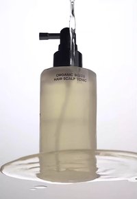 Klar glasflaska med ekologisk hårbotten tonic och svart pumpdispenser. Vätskan inuti är ljusgul, mot en vit bakgrund.