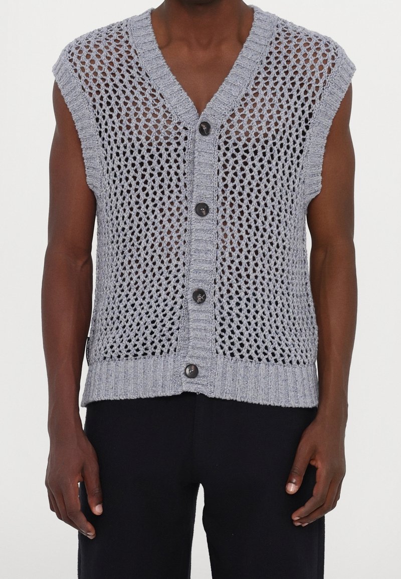 Gilet sans manches gris tricoté avec un grand motif à mailles ouvertes, boutonné sur le devant, porté sur peau nue, assorti à un pantalon noir.
