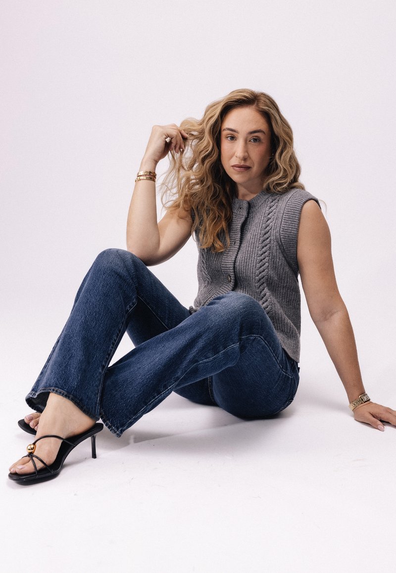 Un gilet en tricot gris à côtes avec des manches courtes, associé à un jean bleu évasé et des talons noirs à lanières avec un accent doré.