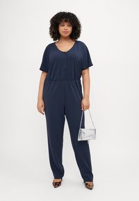 Navy blue jumpsuit with short sleeves, V-neck, button detailing, elastic waist, and loose-fitting legs. Worn with a silver clutch and leopard print heels.