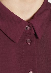 Gros plan d’un col de chemise bordeaux à boutons avec trois boutons visibles et un tissu texturé sur un cou à la peau claire.