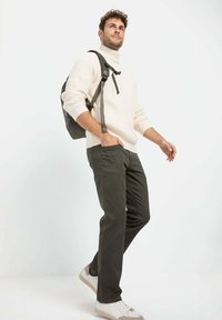 Hellbeige gerippter Rollkragenpullover, dunkelgrüne Hose und weiße Sneakers mit hellen Akzenten. Schwarzer Rucksack mit verstellbaren Trägern.