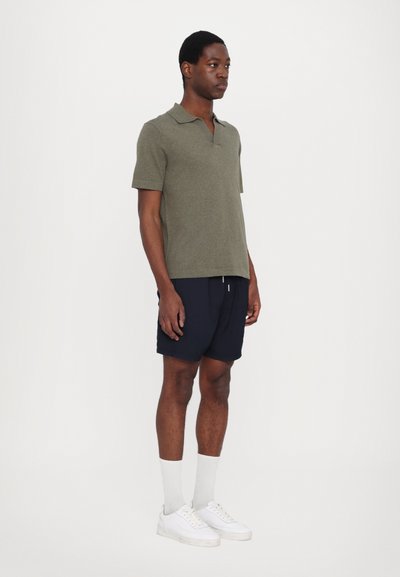 Polo verde-oliva com mangas curtas, combinado com calções azul-marinho. Modelo usa ténis brancos e meias cinzentas claras. Design simples e casual.