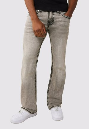 Person trägt verblasste graue Jeans mit dunklen Nähten und weiße Turnschuhe, steht mit einer Hand in der Tasche und der anderen entspannt.