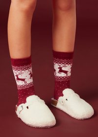 White, fuzzy slippers with a strap detail, paired with red knit socks featuring a reindeer pattern and snowflake accents.