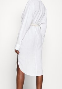Wit linnen jurk met lange mouwen, zijslitten en een tailleband, met een afgeronde zoom en een losse pasvorm. De textuur lijkt licht en ademend.