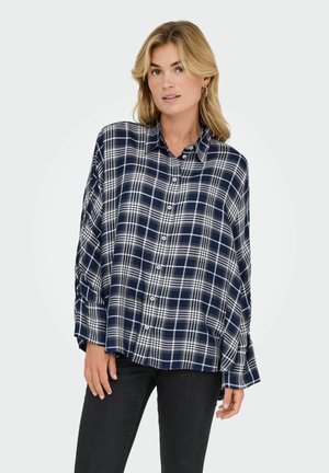 Blonde vrouw in een loszittend marineblauw en wit geruit overhemd met knopen en zwarte broek, poseert met één hand bij haar dij tegen een witte achtergrond.