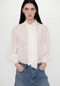 Blusa bianca e trasparente con motivo floreale, maniche lunghe e fiocco al collo. Abbinata a jeans in denim blu a vita alta.