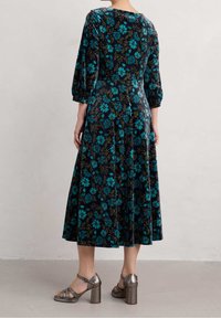 Frau in einem schwarzen Midikleid mit blauem Blumenmuster und taillierter Passform, kombiniert mit metallischen Blockabsatzsandalen, auf einem grauen Boden stehend.