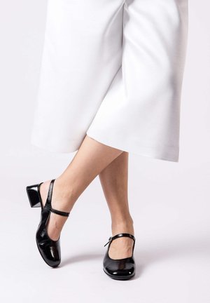Piernas cruzadas con pantalones blancos de pierna ancha y zapatos Mary Jane negros de charol con tacón bajo sobre un fondo claro.