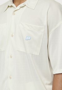Camisa de manga corta, de color beige claro, hecha de una tela suave, que presenta dos bolsillos frontales y un logo 'P' azul claro en el bolsillo izquierdo.