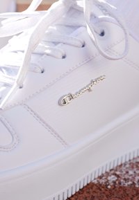 Sneaker bianco realizzato in materiale sintetico liscio con punta rotonda, lacci piatti e logo argento "Champion" sul lato. Suola texturizzata.