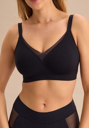 Kobieta w czarnym bezszwowym biustonoszu i dopasowanych majtkach z wysokim stanem z przezroczystymi panelami z siatki na neutralnym tle.