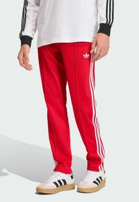 Calças de atletismo vermelhas com riscas brancas nas laterais e um logótipo bordado. Combinadas com sapatilhas brancas com riscas pretas e uma sola texturizada.