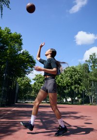 Atletické šortky v tmavosivej farbe s čiernym pásom, skombinované s priliehavým čiernym tričkom a čiernymi teniskami. Basketbalová lopta je vo vzduchu.