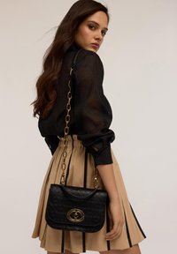 Borsa a mano in faux pelle nera con motivo in rilievo, accessori dorati e tracolla a catena. Abbinata a una gonna beige con righe verticali.