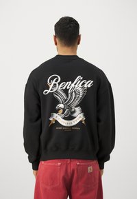 Sudadera negra con un gran gráfico de un águila, texto "Benfica" y una cinta con la frase "E Pluribus Unum". Puños y dobladillo acanalados.