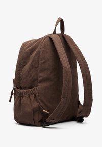 Sac à dos en velours côtelé marron avec deux sangles en tissu, une poche latérale et une forme légèrement arrondie, présentant une texture à côtes verticales.