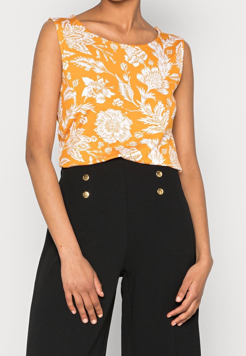 Oranžový květinový crop top s kulatým výstřihem a bezrukávovým designem, v kombinaci s černými kalhotami s vysokým pasem a zlatými knoflíky.