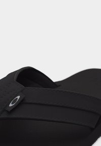 Μαύρη σανδάλι με φαρδιά λουρί που διαθέτει μια μικρή υφασμάτινη ετικέτα με λευκό κυκλικό λογότυπο σε απλό ανοιχτό φόντο.