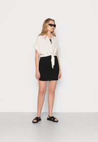 Camisa de botão de manga curta em bege claro, atada na cintura, combinada com uma saia mini preta com franzido e sandálias pretas com tiras cruzadas.